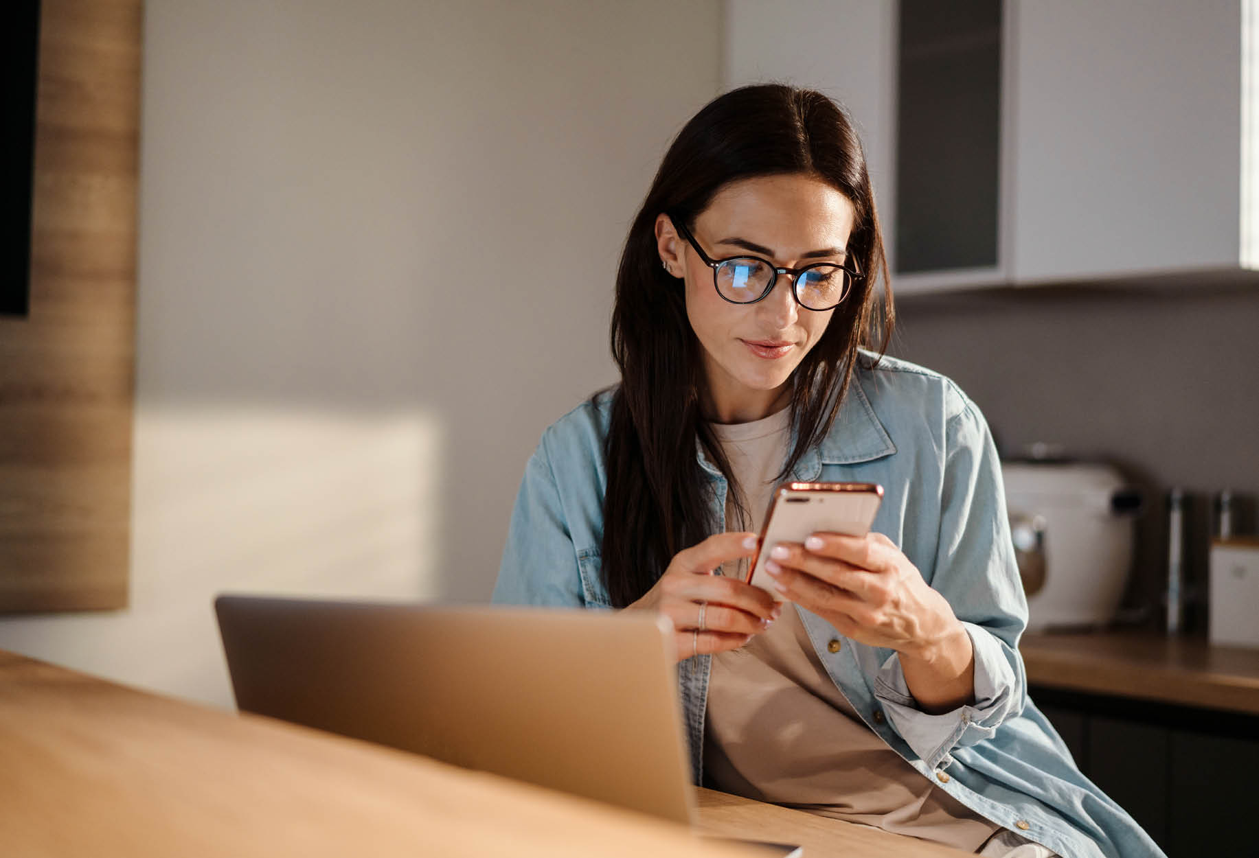 Vrouw zit achter laptop en kijkt op telefoon